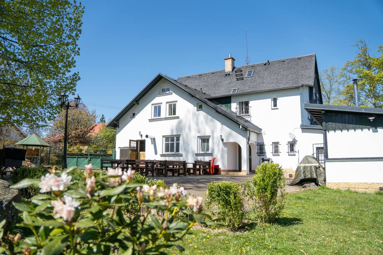 Vlčárna — bílý podstávkový dům s terasou a dlouhými stoly, zahrada s rozkvetlými rododendrony